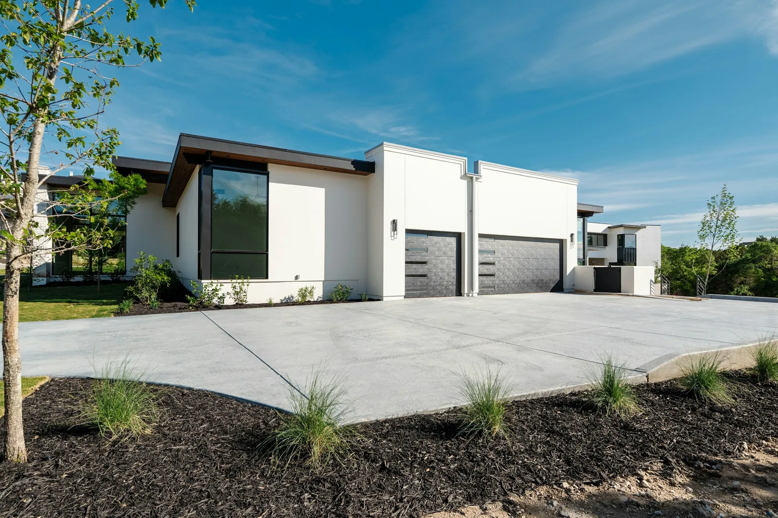 A wide light-gray concrete driveway in front of a modern white house with a two-car garage, with a mulch bed and ornamental grass in front