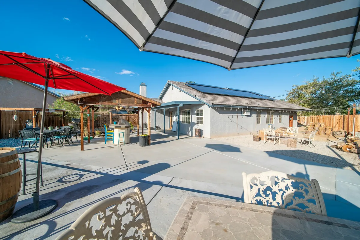 A sunny backyard concrete patio with a striped umbrella overhead, a red umbrella, an outdoor kitchen and seating, next to a single-story house