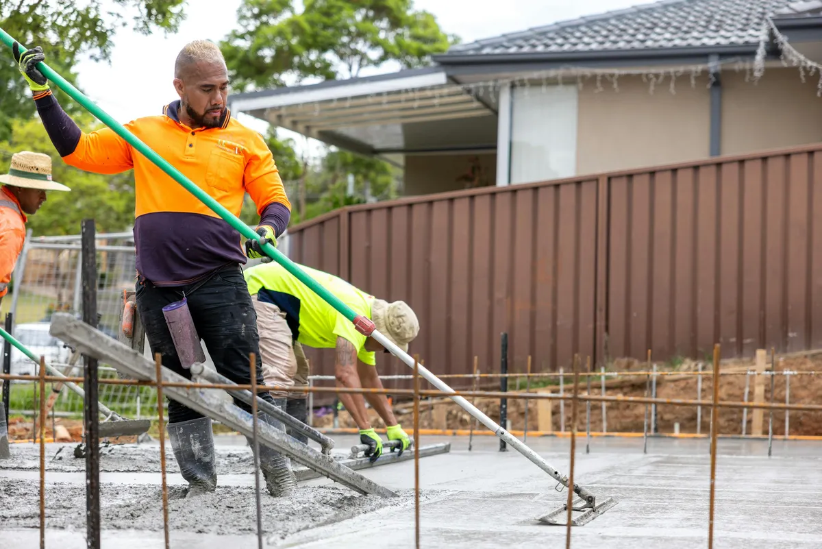 Two workers in orange and yellow hi-vis shirts screeding a fresh concrete slab over rebar, with a brown metal fence and a house behind them