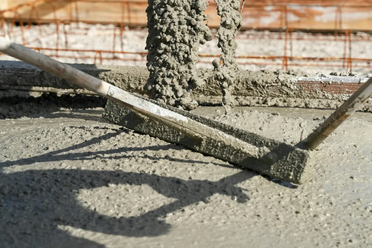 Wet concrete pouring from a chute onto a slab, with a screed board resting in the concrete