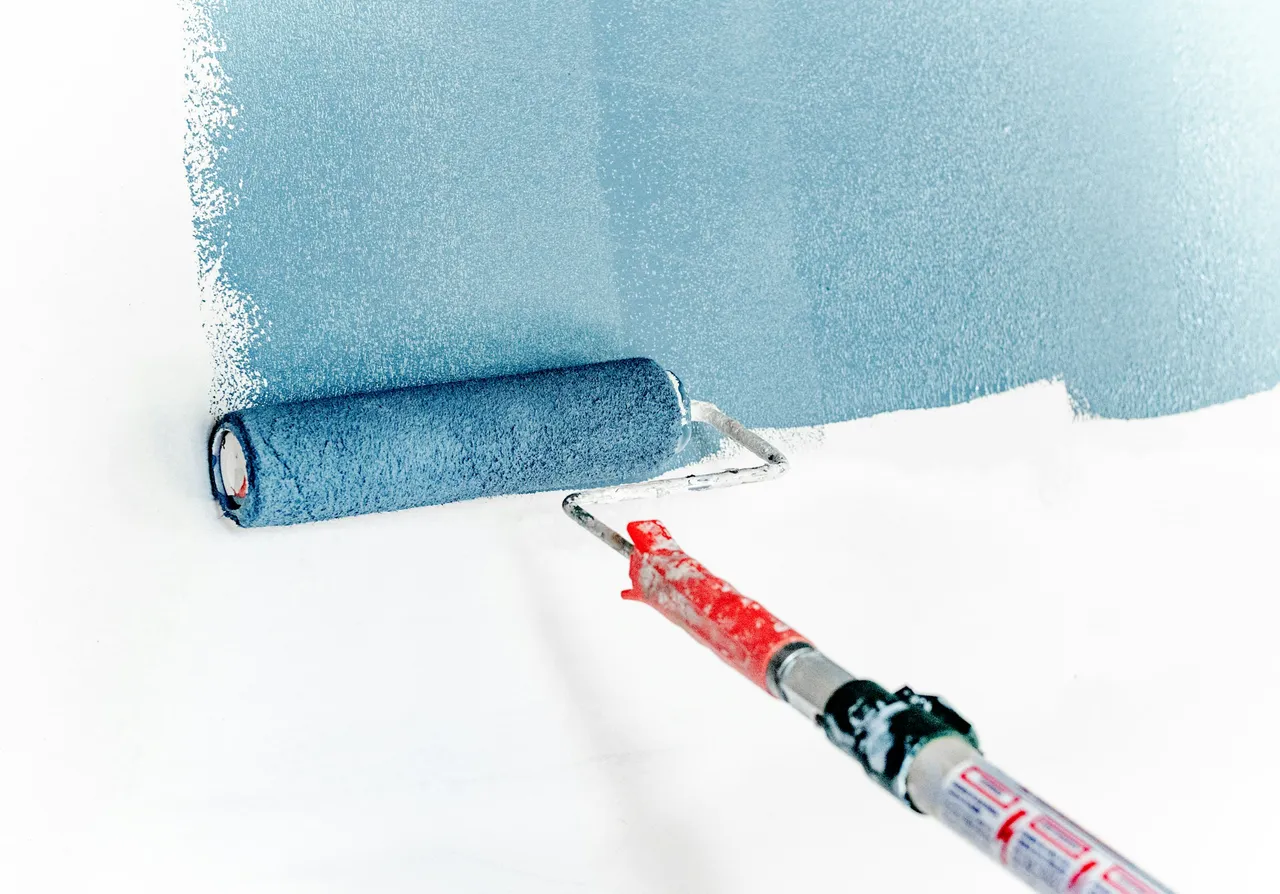 A paint roller spreading pale blue paint over a white wall