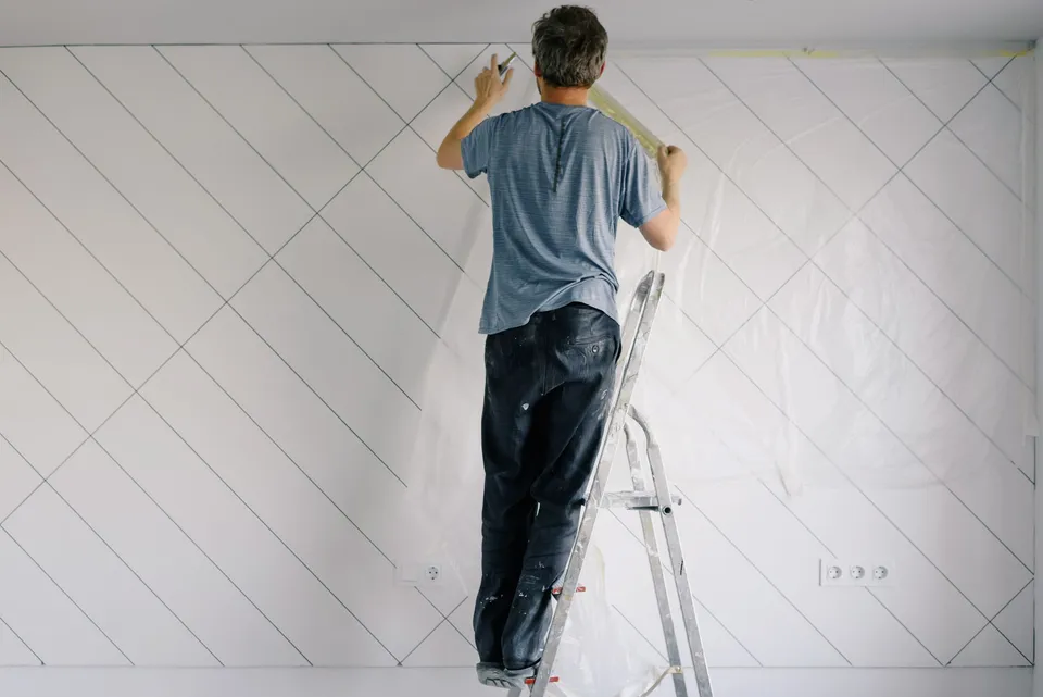 A man on an aluminum stepladder taping plastic sheeting along the top of a white wall before painting