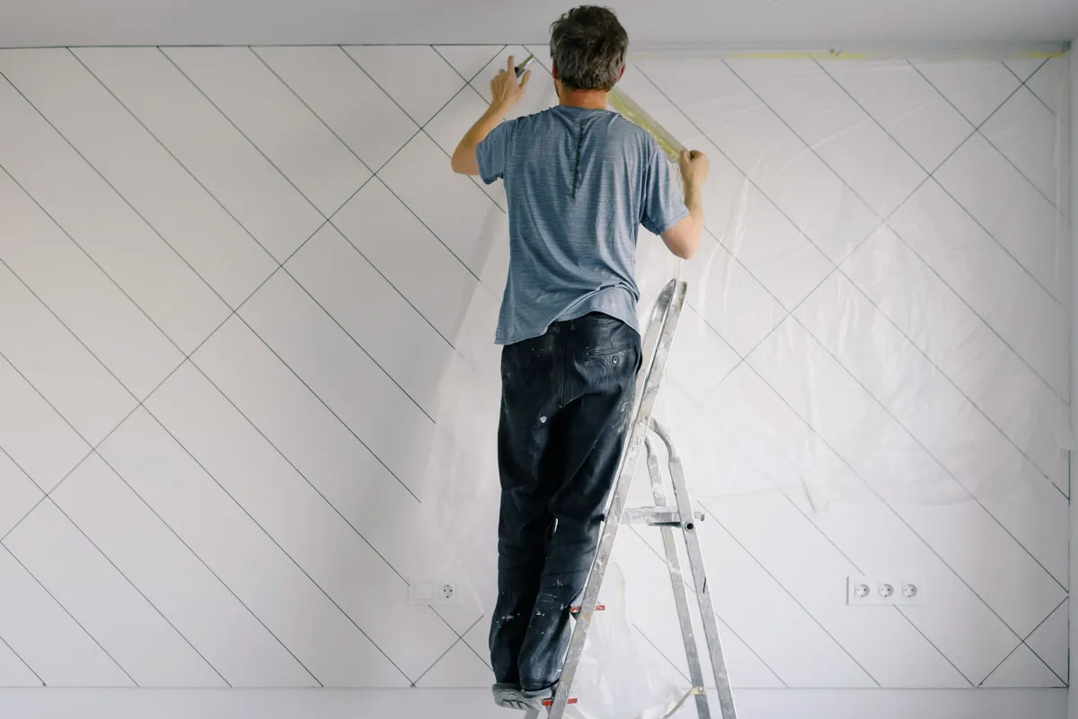 A man on an aluminum stepladder taping plastic sheeting along the top of a white wall before painting